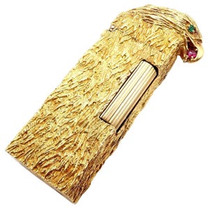 May include: A gold-coloured lighter with an eagle head design. The lighter has a textured surface and is decorated with green and red gemstones for the eyes and beak. The rectangular body of the lighter has a ribbed, gold-coloured metal section.