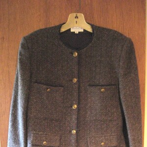 May include: A dark gray tweed blazer with gold buttons and flap pockets. The blazer has a mandarin collar and a slightly cropped length.
