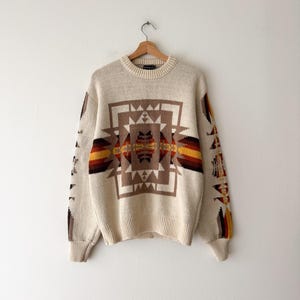 May include: Cream-colored sweater with a geometric pattern in shades of brown, orange, and yellow. The design is centered on the front and repeated on the sleeves. The sweater is hanging on a wooden hanger.