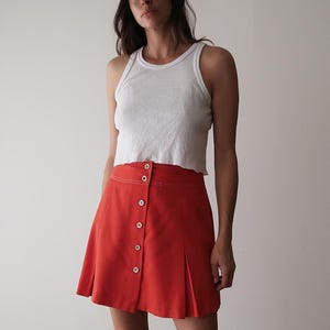 Op de afbeelding: Een vrouw draagt een witte geribbelde tanktop, een rode minirok met knopen en een zwarte baseballcap. De rok heeft witte stiksels en een geplooid ontwerp. De pet heeft een kleine patch.