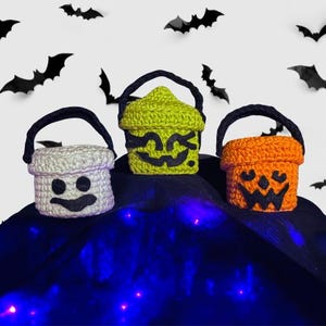 May include: Three crocheted Halloween treat buckets in white, green, and orange, each with a black handle. The white bucket has a ghost face, the green bucket has a jack-o'-lantern face, and the orange bucket has a jack-o'-lantern face. Black bat cutouts are in the background.
