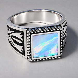 May include: A silver-toned ring featuring a square-shaped inlay with an iridescent blue and white finish. The central square is framed by a twisted rope-like border, and the sides of the band display black recessed floral-inspired engravings.