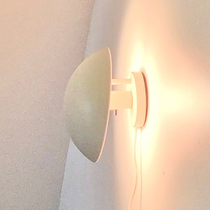 May include: A wall-mounted lamp featuring a matte off-white, dome-shaped shade that directs light toward the wall. The fixture includes a circular base plate and a short, cylindrical arm connecting the shade to the wall, with a power cord extending downward.