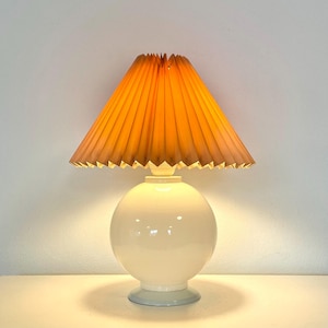 May include: A white ceramic table lamp with a round base and a pleated, light brown lampshade.