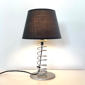 May include: A black lampshade with a silver spring base. The lampshade is made of fabric and has a textured surface. The base is made of metal and has a silver finish.