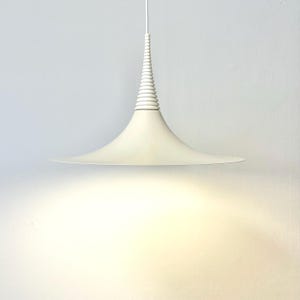 May include: A white pendant light with a wide, flat, circular shade and a tall, narrow, cone-shaped center section. The light is suspended from the ceiling by a thin cord.