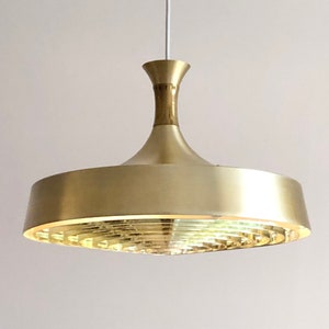 May include: A mid-century modern pendant light fixture with a gold finish. The light has a flared, disc-shaped shade with a series of concentric rings at the bottom. The fixture is suspended from a thin wire and has a brass-colored stem.