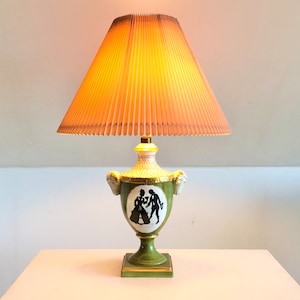May include: A decorative table lamp with a pleated lampshade. The lamp base is green with gold accents and a black and white silhouette design. The lampshade is a warm, light orange color.