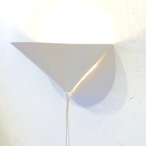 May include: A white, triangular wall sconce with a minimalist design. The sconce is mounted on a textured white wall and has a cord hanging down. The light source is not visible, but the sconce casts a soft glow.