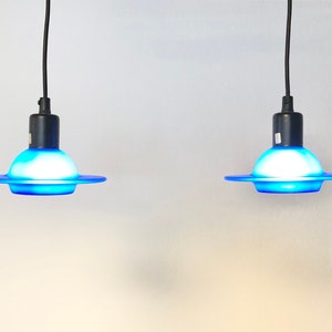 May include: Two blue glass pendant lights with white frosted glass shades hanging from black cords against a white wall.