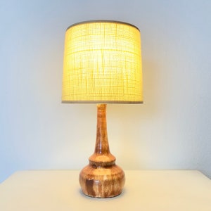 Puede incluir: Una lámpara de mesa de cerámica con un esmalte marrón y beige. La lámpara tiene una base redonda y bulbosa y un cuello cilíndrico. La pantalla es de tela beige claro con una textura tejida.
