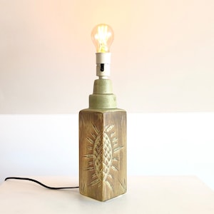 Könnte beinhalten: Eine grüne Keramik-Tischlampe mit einem geschnitzten Sonnenmotiv. Die Lampe hat eine weiße Fassung und eine eingeschaltete Glühbirne.