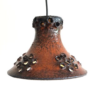 May include: A brown ceramic pendant light with a textured finish and cut-out details. The light has a bell shape and is suspended from a black cord.