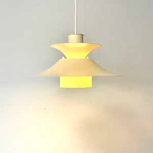 May include: Cream-colored pendant light fixture with a tiered, saucer-like shade design. The light is suspended from a white cord and emits a warm, yellow glow. The fixture has a mid-century modern aesthetic.