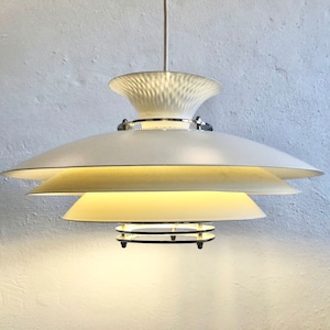 May include: A white, layered pendant light fixture with a textured top and a series of descending, saucer-shaped shades. The light fixture has a chrome accent and emits a warm glow. The design is modern and minimalist.