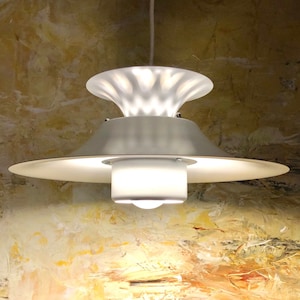 May include: A modern, white pendant light fixture with a layered design. The light has a wide, circular shade and a smaller, textured top section. The fixture is suspended from a white cord and emits a soft, diffused glow.