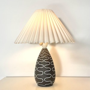 Könnte beinhalten: Eine Keramik-Tischlampe mit einem weißen, gefalteten Lampenschirm. Die Lampenbasis ist braun mit einem weißen, abstrakten Muster.