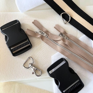 Puede incluir: Un surtido de suministros de costura y manualidades, que incluye hebillas de pl&aacute;stico negro, un cierre de metal plateado, cremalleras beige y una correa a rayas con un anillo de metal plateado. Los art&iacute;culos est&aacute;n dispuestos sobre una tela clara.