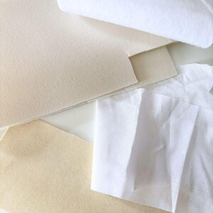 Puede incluir: Vista cenital de varias muestras de tela en tonos blancos y beige. Las telas incluyen una pieza enrollada, cuadrados doblados y piezas cortadas, mostrando diferentes texturas y grosores. La disposici&oacute;n sugiere un enfoque en textiles y materiales.
