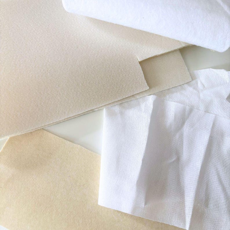 Puede incluir: Vista cenital de varias muestras de tela en tonos blancos y beige. Las telas incluyen una pieza enrollada, cuadrados doblados y piezas cortadas, mostrando diferentes texturas y grosores. La disposici&oacute;n sugiere un enfoque en textiles y materiales.