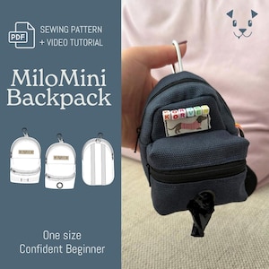 May include: A dark blue mini backpack with a silver carabiner clip. The front features a small rectangular patch with the text KORVEN and a graphic of a dachshund. A black plastic bag dispenser is attached to the bottom zipper compartment.