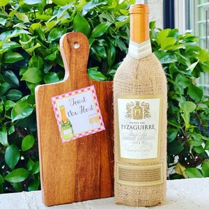 May include: A wooden cutting board with a pink and white card that says "Toast the Host" and a bottle of wine wrapped in burlap. The wine bottle label says "Vino de Eyzaguirre Cabernet Sauvignon 2013 Produce Chile."