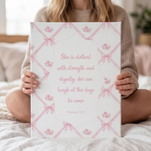 May include: A white canvas print featuring a pink ribbon and floral design. The text reads, "She is clothed with strength and dignity, she can laugh at the days to come." The print is held by a person sitting on a bed.