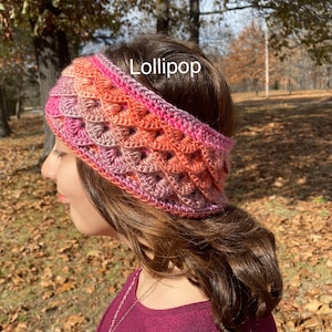 May include: A crocheted headband in shades of pink, orange, and peach. The headband has a scalloped edge and is made with a textured stitch. The headband is worn by a person with long brown hair. The text "Lollipop" is visible on the headband.