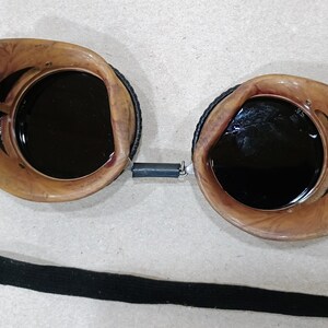 May include: Vintage-style goggles with dark lenses and a brown, marbled frame. The goggles have a black elastic strap for securing them and a spring connecting the lenses. These protective glasses are suitable for various activities.