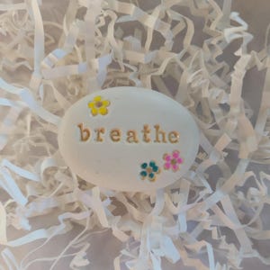 May include: White painted stone with the word "breathe" in gold lettering and colorful flower accents.