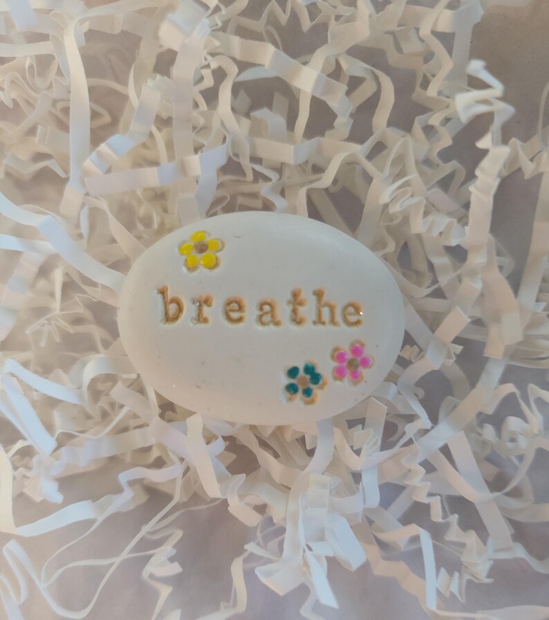 May include: White painted stone with the word "breathe" in gold lettering and colorful flower accents.