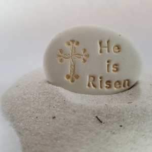 May include: White stone with a gold cross and the words "He is Risen" engraved on it. The stone is sitting on a bed of white sand.