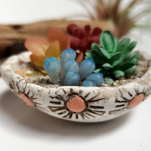 May include: A small white ceramic planter with a textured surface and pink sun designs. The planter is filled with artificial succulents in shades of blue, green, and orange.
