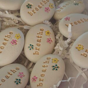 May include: White painted stones with the word "breathe" written in gold lettering. Each stone has colorful flowers painted on them.