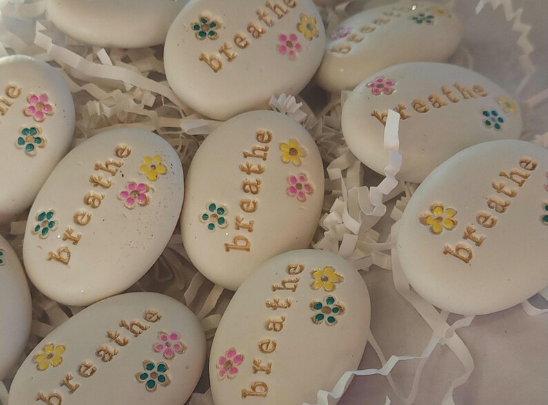 May include: White painted stones with the word "breathe" written in gold lettering. Each stone has colorful flowers painted on them.