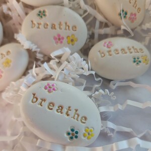May include: White painted stones with the word "breathe" written on them. Each stone has colorful flowers painted on them.