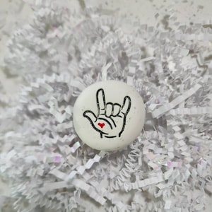 May include: White round ceramic coaster with a black and white illustration of the American Sign Language symbol for "I love you". The sign has a small red heart in the palm of the hand.