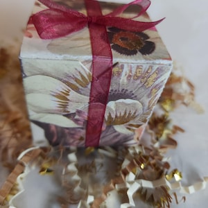May include: A small, square gift box with a floral design and a burgundy ribbon tied in a bow. The box is sitting on a bed of shredded paper.