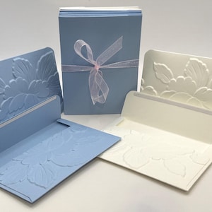 May include: A collection of stationery featuring embossed floral designs. The set includes light blue and off-white cards and envelopes. A stack of cards is tied with a sheer ribbon. The cards have a detailed floral pattern.