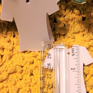 May include: A stack of white t-shirt shaped gift tags with a hole punched at the top. The tags are measured against a ruler, showing they are approximately 5 cm tall.