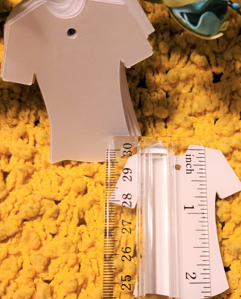 May include: A stack of white t-shirt shaped gift tags with a hole punched at the top. The tags are measured against a ruler, showing they are approximately 5 cm tall.