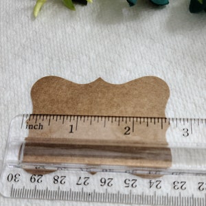 K&ouml;nnte beinhalten: Ein braunes Papieretikett mit gewelltem Rand hinter einem durchsichtigen Kunststofflineal. Das Lineal zeigt, dass die Breite des Etiketts etwa 6,35 cm betr&auml;gt. Das Etikett hat eine glatte, matte Oberfl&auml;che und eine symmetrische, dekorative Form.