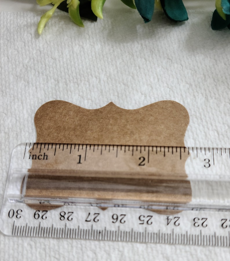 K&ouml;nnte beinhalten: Ein braunes Papieretikett mit gewelltem Rand hinter einem durchsichtigen Kunststofflineal. Das Lineal zeigt, dass die Breite des Etiketts etwa 6,35 cm betr&auml;gt. Das Etikett hat eine glatte, matte Oberfl&auml;che und eine symmetrische, dekorative Form.