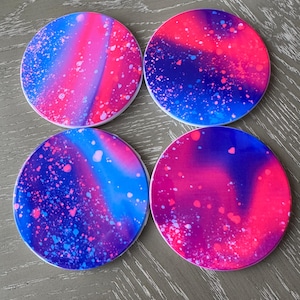 May include: Set of four round coasters with a pink, blue, and white galaxy-inspired design. The coasters have a white background with a swirling pattern of pink, blue, and white paint splatters.