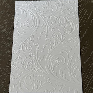 May include: A white rectangular card with an embossed floral design. The pattern includes swirling vines, leaves, and dotted details, giving a textured, elegant look. The card is clean and crisp, with a slight sheen.