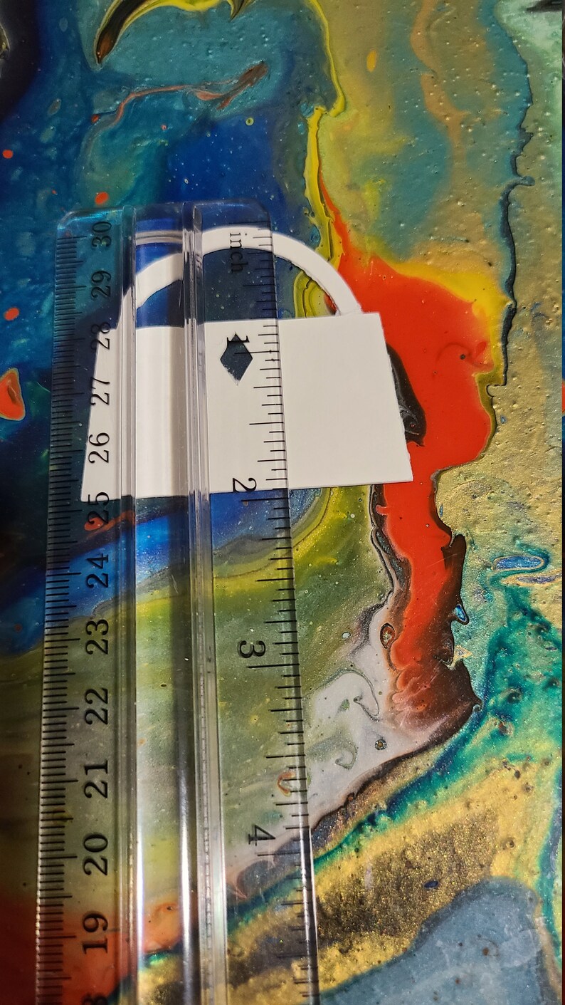 May include: A white paper lock shape with a black diamond in the center, measured with a ruler in inches. The lock is approximately 2 inches tall and 1.5 inches wide.