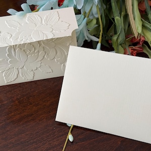 Op de afbeelding: Ivoor briefpapier set met een reliëf bloemenontwerp. De set bevat een envelop met een gedetailleerd bloemenpatroon en een blanco kaart. Het briefpapier ligt op een donkere houten ondergrond, met bloemaccenten op de achtergrond.