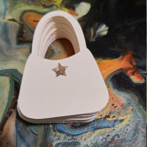 May include: A stack of white paper cutouts shaped like a handbag with a star cutout in the center.