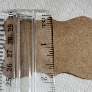 K&ouml;nnte beinhalten: Ein beigefarbenes Papieretikett mit gewelltem Rand unter einem transparenten Kunststofflineal. Das Lineal zeigt, dass die Breite des Etiketts etwa 3,8 cm betr&auml;gt. Das Etikett hat eine glatte, matte Textur und eine hellbraune Farbe.