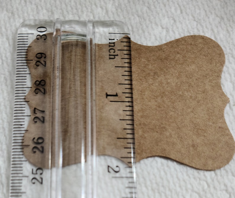 K&ouml;nnte beinhalten: Ein beigefarbenes Papieretikett mit gewelltem Rand unter einem transparenten Kunststofflineal. Das Lineal zeigt, dass die Breite des Etiketts etwa 3,8 cm betr&auml;gt. Das Etikett hat eine glatte, matte Textur und eine hellbraune Farbe.